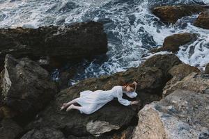Barefoot woman in long white dress wet hair lying on a rocky cliff vacation concept photo
