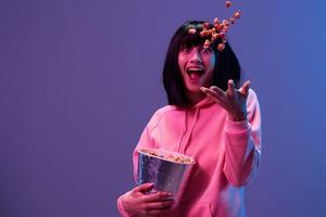 Crazy excited awesome brunet woman in pink hoodie throws up popcorn open mouth posing isolated in blue violet color light studio background. Neon party Cyberpunk Cinema concept. Copy space photo