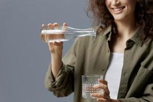 Happy smiling curly beautiful woman in casual khaki green shirt pours water from bottle in glass posing isolated on over gray blue background. Healthy lifestyle. Water is Life concept. Free place photo