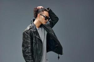 Side of view of stylish tanned curly man leather jacket trendy sunglasses posing isolated on gray studio background. Cool fashion offer. Huge Seasonal Sale New Collection concept. Copy space for ad photo