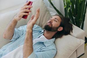 un hombre con un barba mentiras en el sofá durante el día a hogar y mira a su teléfono relajante en su día apagado, un hombre juego en el valores mercado en línea en su teléfono foto