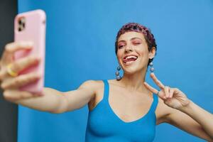 Young sports woman fashion blogger takes a picture of herself on the phone in blue sportswear smiling and showing her tongue on a blue monochrome background photo