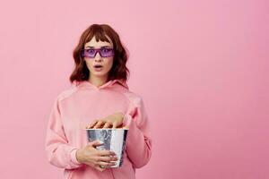 Cool movie. Confused cute redhead lady in pink hoodie sunglasses with popcorn posing isolated on over pink studio background show copy space Banner. Fashion Cinema concept. Entertainment offer photo