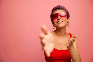 Moda retrato de un mujer con un corto Corte de pelo en de colores Gafas de sol con raro accesorios con pendientes sonrisas en un rosado brillante antecedentes muestra su manos a el cámara foto