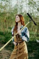 A woman farmer in work clothes and an apron works outdoors in nature and holds a rake to gather grass photo