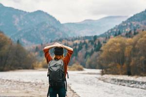 happy woman hiker with a backpack on the river bank look into the mountains and autumn forest nature photo