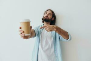 Lanza libre milenario hombre con un barba Bebiendo café desde un reciclado taza en elegante hipster ropa blanco camiseta azul pantalones y camisa en un blanco antecedentes foto