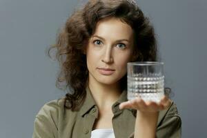 Serious adorable curly beautiful woman in casual khaki green shirt holds suggests glass of water posing isolated on over gray blue background. Healthy lifestyle. Water is Life concept. Copy space photo