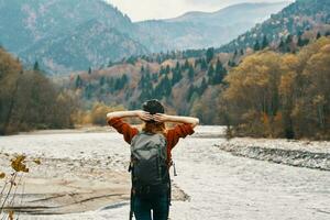 a traveler in a sweater hat with a backpack gestures with her hands and relaxes on the river bank in the mountains photo