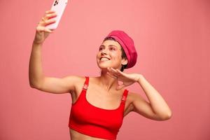 A young woman blogger with colored pink hair and a short haircut takes a picture of herself on the phone and broadcasts a smile in stylish clothes and a hat on a pink background monochrome style photo