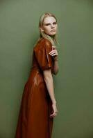 Handsome pretty blonde stylish young lady in brown palette leather dress serious looking over her shoulder at camera posing isolated on over olive green studio wall background. Fashion portrait banner photo