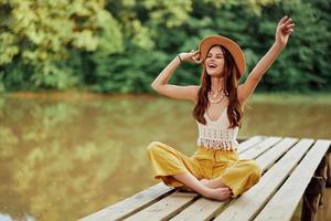 A young hippie woman sits on a lake bridge wearing stylish eco clothes and smiling photo