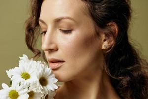 Close up portrait of cheerful curly female in linen casual shirt hold chamomiles enjoy flowers smell close eyes posing isolated on olive green pastel background. Nature is beauty concept. Copy space photo