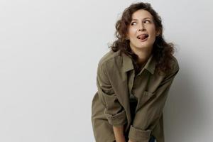 Cheerful happy curly beautiful woman in casual khaki green shirt searches with tongue looks aside enjoy cool day posing isolated on over white background. People Emotions Lifestyle concept. Copy space photo