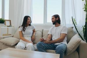 A man and a woman sitting at home on the couch in white stylish t-shirts and chatting merrily smiling and laughing at home. Male and female friendship photo