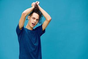 Positive joyful curly tanned Caucasian guy in basic t-shirt touches plays with hair curls posing isolated on over blue background. Lifestyle and Emotions concept. Good offer with copy space for ad photo