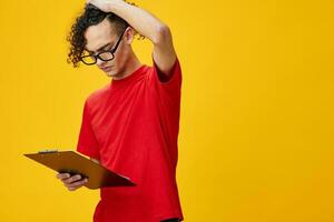Thoughtful myopic young student man in red t-shirt funny eyewear holds tablet folder with study notes posing isolated on over yellow studio background. Free place for ad. Education College concept photo