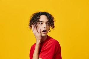 Shocked confused funny Caucasian young man in red t-shirt touches cheeks posing isolated on over yellow studio background. The best offer with free place for advertising. Emotions for everyday concept photo