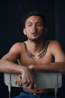 Modelling snapshots. Pensive serious tanned attractive handsome naked man sit on chair smoking looks aside posing isolated in black studio background. Fashion offer. Copy space for ad. Closeup photo