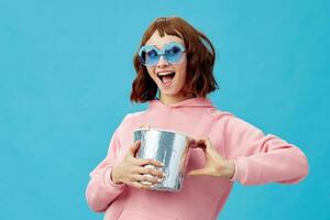 Movie concept. Happy cheerful cute redhead lady in pink hoodie sunglasses with popcorn posing isolated on blue studio background. Copy space Banner. Fashion Cinema. Entertainment offer photo