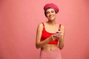 joven mujer blogger con de colores rosado pelo y corto Corte de pelo voltear mediante el teléfono pantalla y mecanografía un mensaje con un sonrisa y sorpresa en elegante ropa en un rosado antecedentes foto