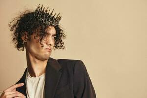 Dissatisfied handsome stylish funny narcissistic tanned curly man with crown posing isolated on over beige pastel background. Fashion New Collection offer. Retro style concept. Free place for ad photo