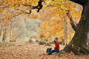 mujer en otoño en el parque cerca un grande árbol y en un mochila en el suelo foto
