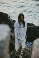 bonito mujer en blanco vestir soportes en el rocas Oceano naturaleza libertad foto
