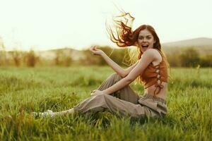 un mujer se sienta en el verde joven Fresco césped en el abierto aire y se ríe y sonrisas felizmente en el puesta de sol luz, su volador rojo pelo revoloteando en el viento foto