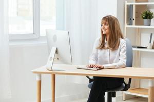 Cheerful happy adorable blonde businesswoman worker freelancer look at screen work on desktop computer online in light modern office. Happy employee writes quarterly report. Copy space, remote job photo