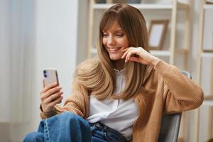 Cute young blonde lady in warm sweater doing video call with friends use phone sitting in armchair at modern home interior. Pause from work, take a break, social media in free time concept. Copy space photo