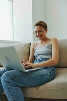 Teenage girl freelancer with laptop sitting on couch at home smiling in home clothes and glasses with short haircut, lifestyle with no filters, free copy space photo