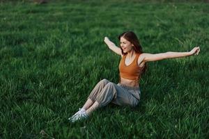 A young redheaded woman sits on the green grass in the park with her arms outstretched in different directions and smiling in the light of the setting sun photo