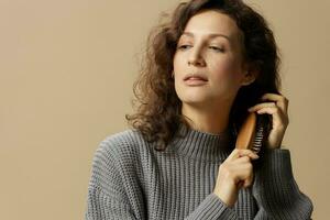 Calm pretty curly beautiful female in gray casual sweater with hairbrush combs hair looks aside posing isolated on over beige pastel background. Problematic unruly damaged hair concept. Copy space photo