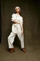 fashionable woman in hat jumpsuit and brown shoes lifted her leg up on brown background photo
