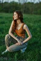 A beautiful redheaded woman sits on the grass in sporty summer clothes and relaxes after a walk in nature and looks out at the sunset. photo