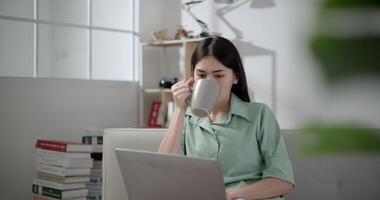 Mano disparo, asiático joven mujer sentado y bebida bebida en taza, ella sonrisa con contento mientras utilizar ordenador portátil computadora en vivo habitación a hogar video