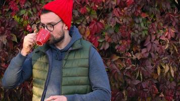 Mann im ein rot Deckel mit ein Tasse im seine Hände Trinken Tee oder Kaffee draußen. Kaffee Unterbrechung. video