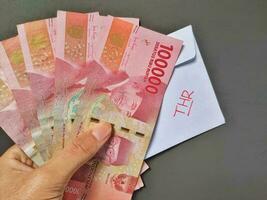 Man holding a white envelope written of THR and new Indonesian banknotes, usually Tunjangan Hari Raya or called THR are given to employees ahead of Eid. Isolated on black background and top view photo