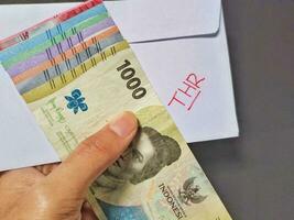 Man holding a white envelope written of THR and new Indonesian banknotes, usually Tunjangan Hari Raya or called THR are given to employees ahead of Eid. Isolated on black background and top view photo