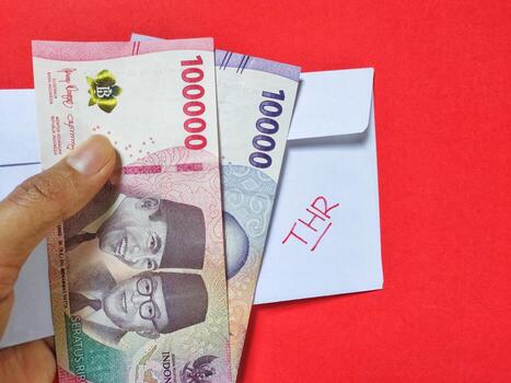 Man holding a white envelope written of THR and new Indonesian banknotes, usually Tunjangan Hari Raya or called THR are given to employees ahead of Eid. Isolated on red background and top view. photo