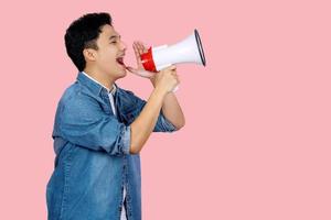 Happy young Asian man in blue shirt shouting announce into megaphone isolated on pink background in studio. photo