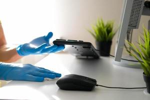 Male hands in rubber gloves wipe the white table and keyboard from the computer. Disinfection in the office during the COVID-19 pandemic. photo