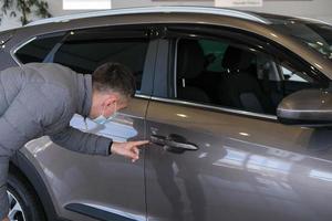 un hombre en un médico máscara examina un coche en el cabina para defectos coche alquilar. comprando un coche foto