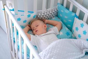 A charming boy sleeps in a baby white crib. A small child sleeps in a crib on blue bedding. Sleep workout concept. photo