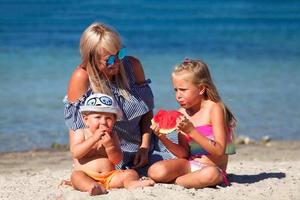 Mom and children are sitting on the beach near the sea and eating watermelon. Vacation. photo