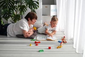 Grandmother is lying on the floor with her grandson and watching a book. The nanny is engaged in developing tasks with a toddler boy. Early development. photo