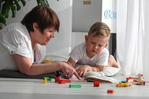 Grandmother is lying on the floor with her grandson and watching a book. The nanny is engaged in developing tasks with a toddler boy. Early development. photo