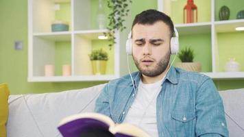 contento Universidad estudiante leyendo un libro a hogar escuchando a música. joven estudiante hombre leyendo un libro a hogar solo escuchando a música con auriculares. video