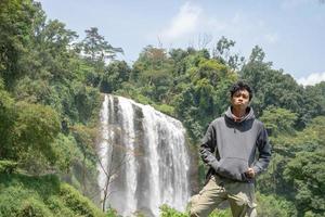 Man stand wear hoody in front of the great water fall with hand reel, Semarang central Java. The photo is suitable to use for adventure content media, nature poster and forest background.